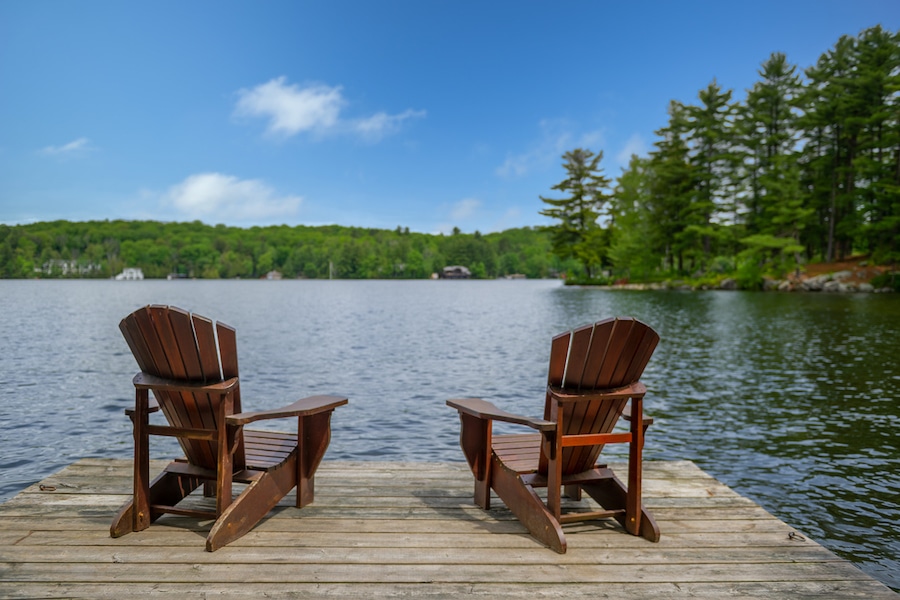 Two Adirondack chairs on a wooden dock facing a lake in Muskoka, Ontario Canada during a sunny summer morning. Cottages are nested between trees across the water