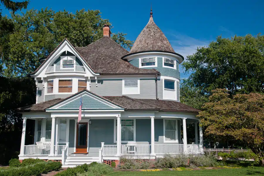 Beautiful gray traditional victorian house.  House has an American Flag haning over the porch and shows a beautiful garden with flowers and trees.  Set against a cloudless blue sky