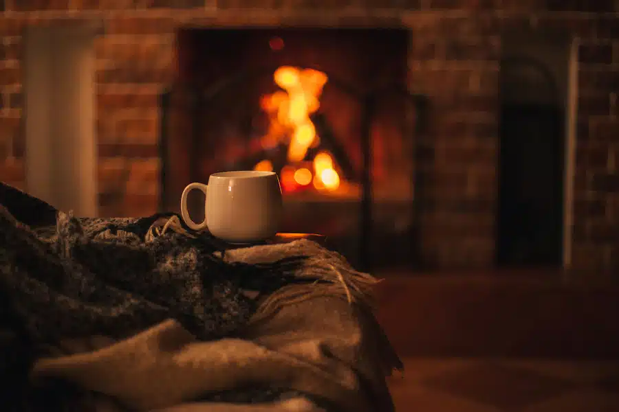Mug with hot tea standing on a chair with woolen blanket in a cozy living room with fireplace. Cozy winter day.