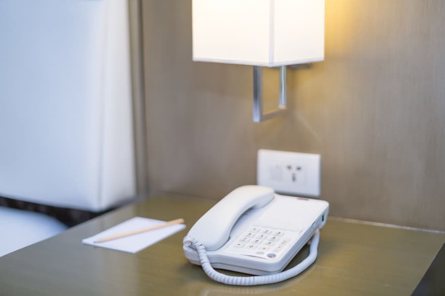 Hotel bedroom white Telephone on a bedside table with note pad and pencil a bedside lamp and plug socket all in a bright scene from lamp lightning