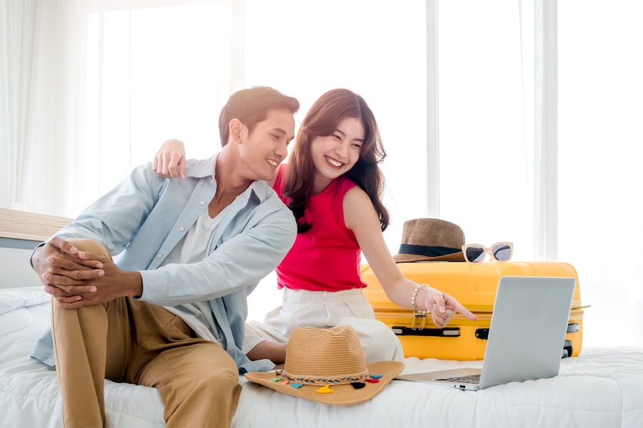 Preparing trip and vacation plan concepts. Young pretty Asian woman and man in denim shirt looking at the laptop and map on the bed with suitcase in bedroom. Travel planning on summer holiday.