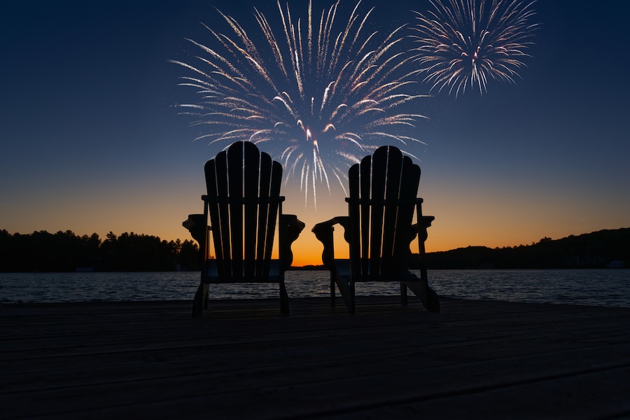 Fireworks over two Adirondack chairs on the wooden dock facing the sunset orange hues of sunset over the calm water.
