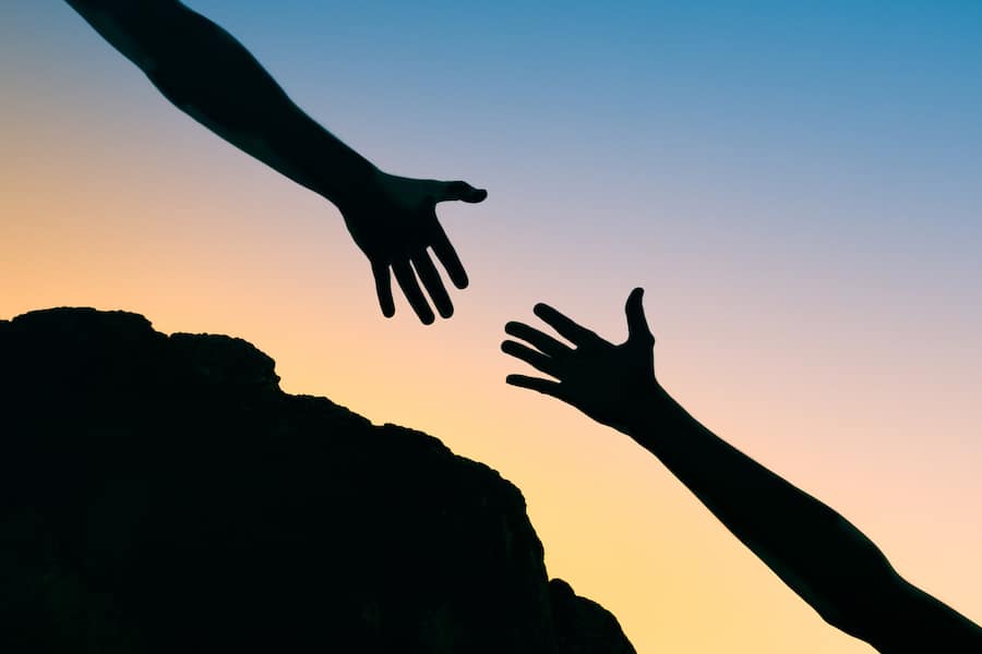 Persons hand reaching out to help another hand up a mountain cliff.