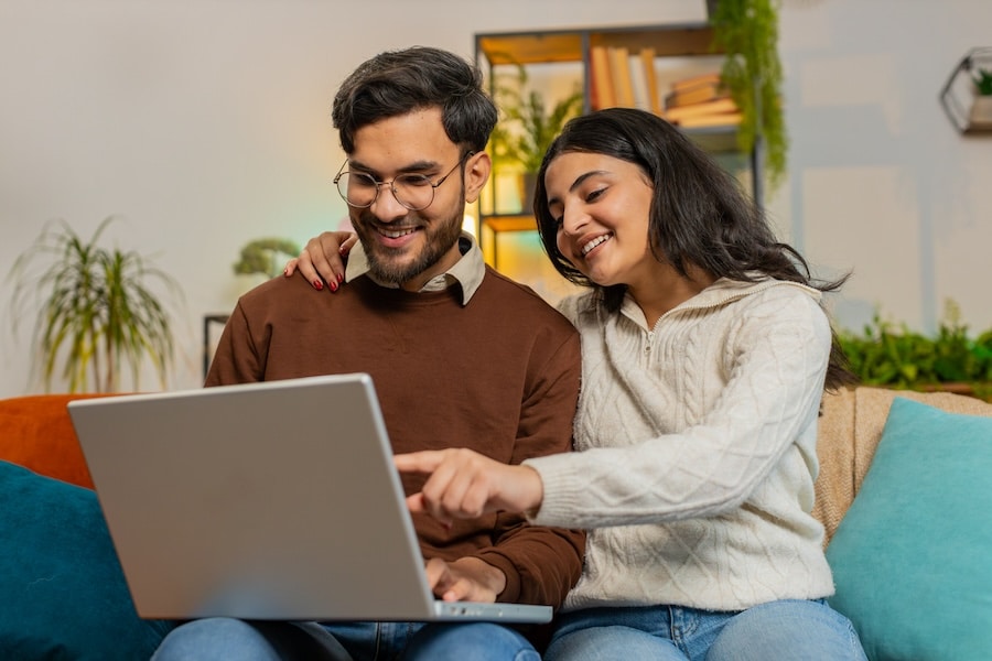 Cheerful young diverse couple in casual clothes discussing travel plans while using laptop on sofa in living room at home.