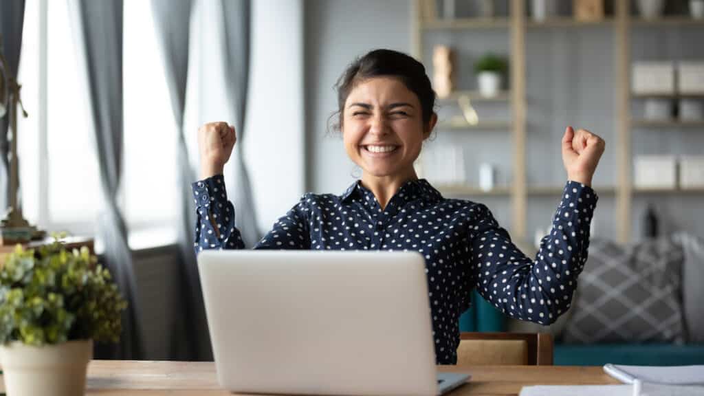 Euphoric young indian girl student winner celebrate victory triumph sit at home desk after managing to reduce the level of spam in her email inbox